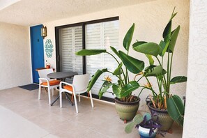 Eine Terrasse mit Tisch und Stühlen, Kübelpflanzen, einer blauen Tür und einem Fenster mit Jalousien.