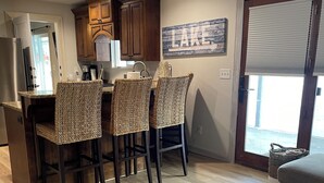 A kitchen with wooden cabinets, a dining area with wicker chairs, a coffee maker, and a 'LAKE' sign on the wall.