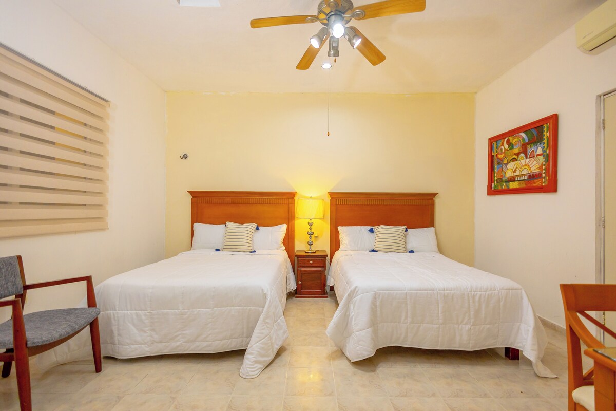 Habitación de hotel con dos camas, un ventilador de techo, una mesita de noche con lámpara y un cuadro en la pared.