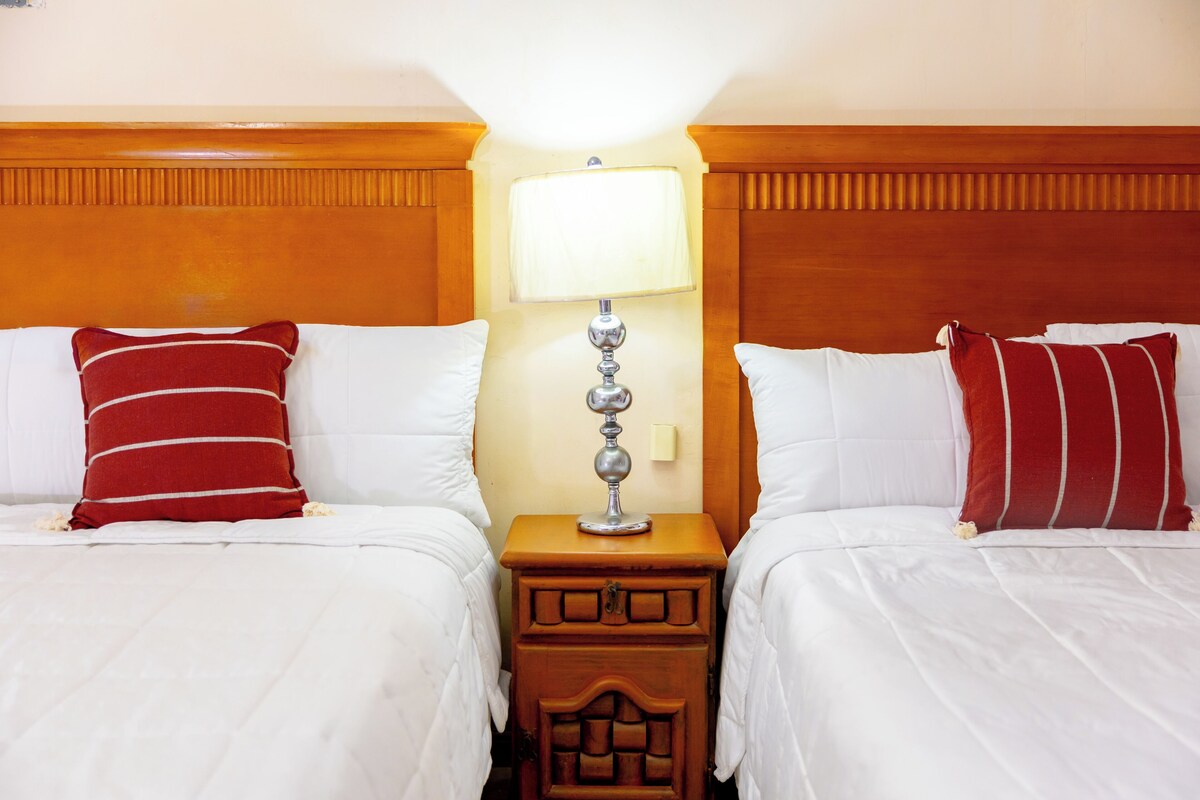 Una habitación de hotel con dos camas, una mesita de noche con lámpara y un cabecero de madera.