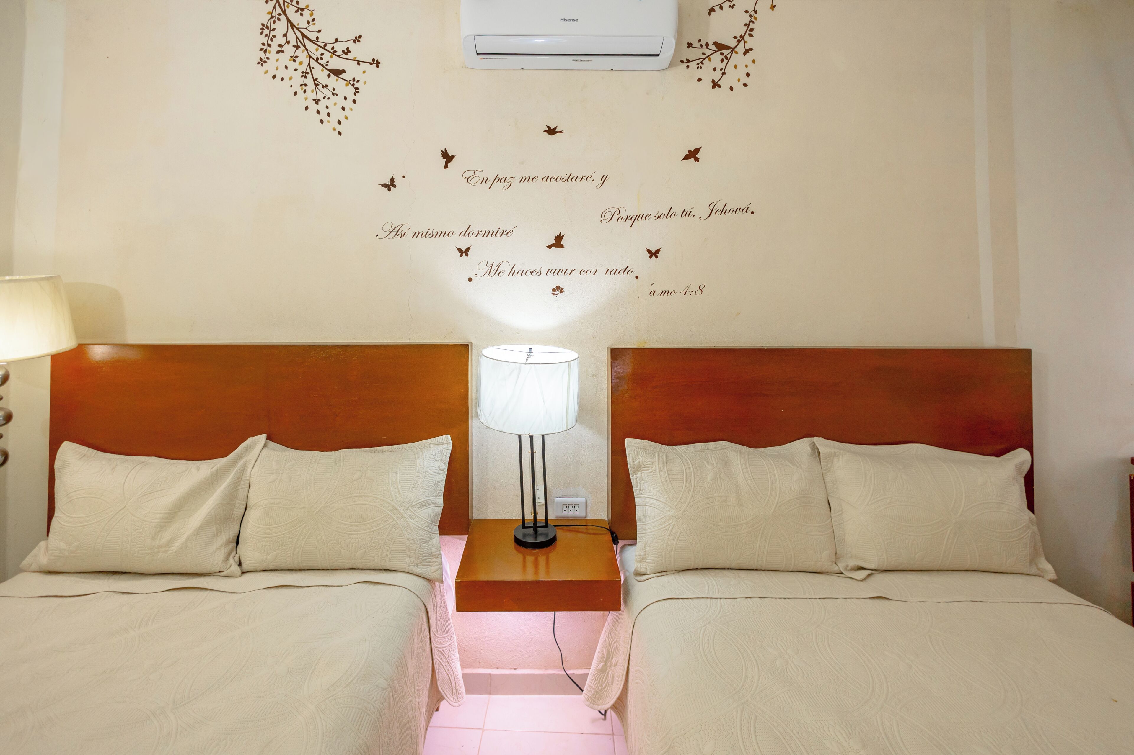 Una habitació d'hotel amb dos llits, una tauleta de nit amb una làmpada i una decoració mural amb text i papallones.