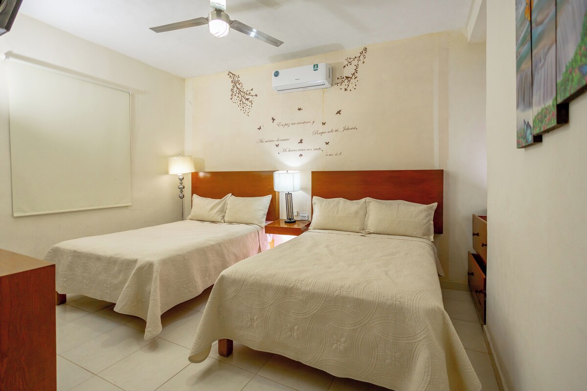 Habitación de hotel con dos camas, ventilador de techo, aire acondicionado, una decoración de pared con texto y una mesita con lámpara.