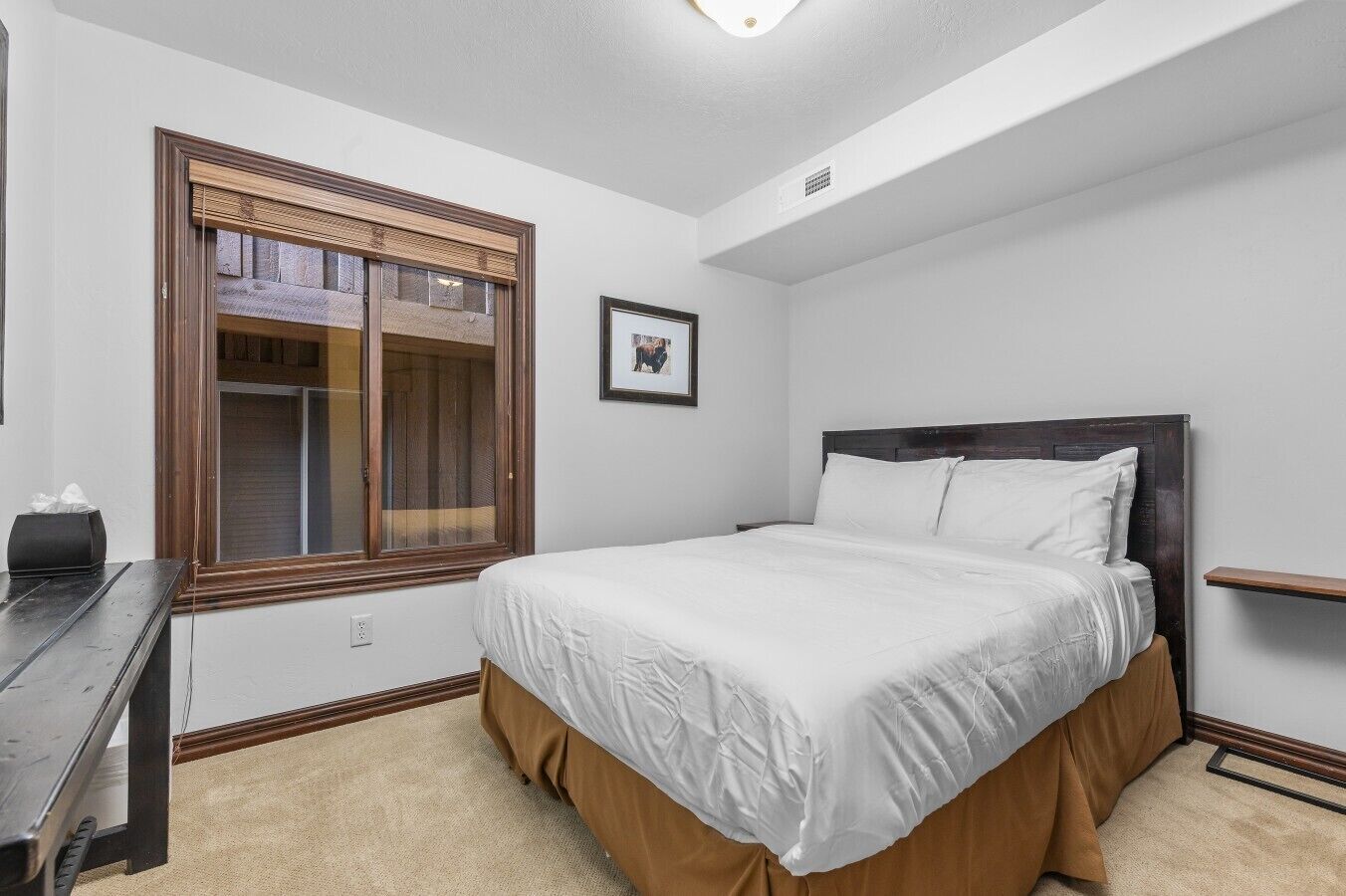 A bedroom with a bed, a window with blinds, a picture on the wall, and a desk with a lamp.