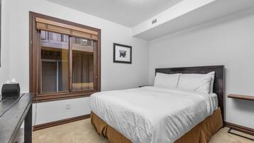 A bedroom with a bed, a window with blinds, a picture on the wall, and a desk with a lamp.