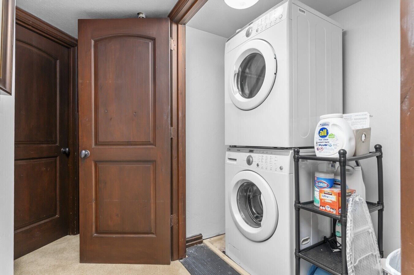 A laundry room with two stacked front-loading washing machines, a shelf with cleaning supplies, and a wooden door.