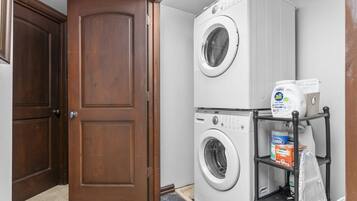 A laundry room with two stacked front-loading washing machines, a shelf with cleaning supplies, and a wooden door.