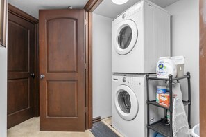 Un lavadero con dos lavadoras de carga frontal apiladas, un estante con productos de limpieza y una puerta de madera.