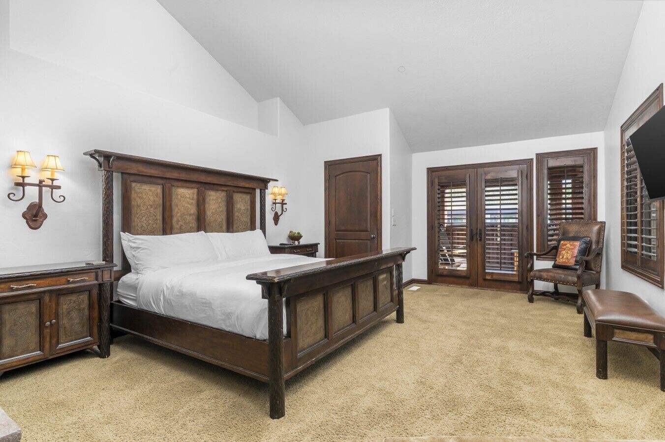 A bedroom with a large wooden bed, two bedside tables with lamps, a chair, and a door with blinds.