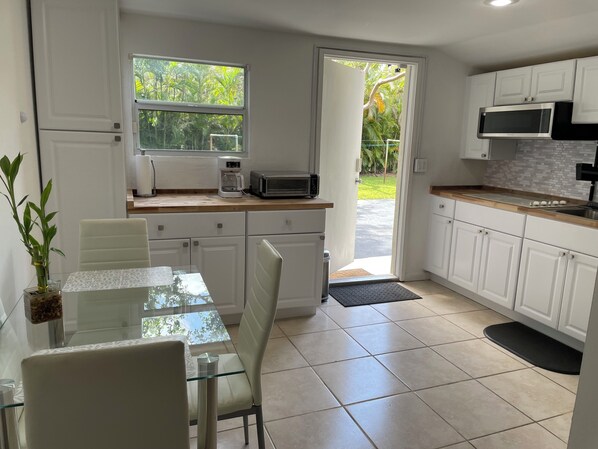 A modern kitchen with white cabinets, a glass dining table, and a view of greenery outside.
