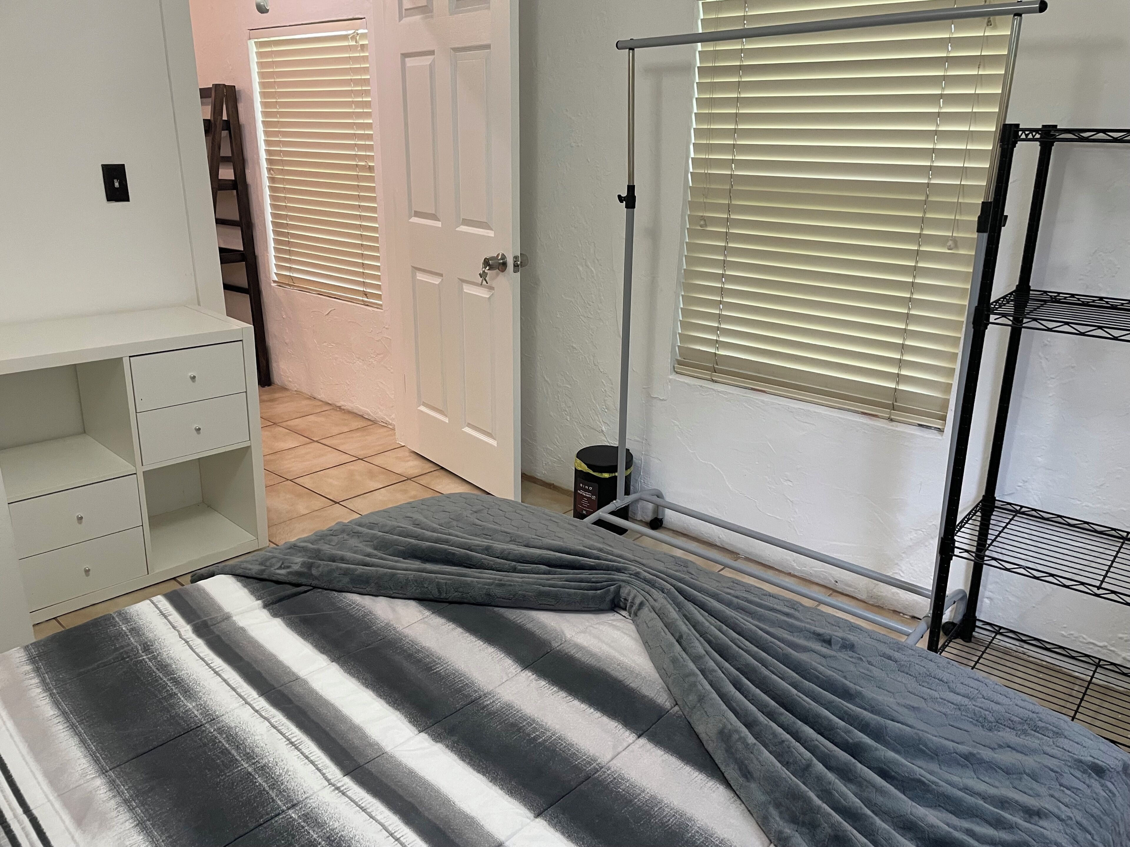 Un dormitorio con cama, armario, radiador, ventana con persianas y puerta.