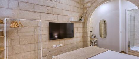 Une chambre avec un mur en briques, un téléviseur à écran plat, un lit recouvert d’une couverture blanche, une étagère en bois et un miroir.