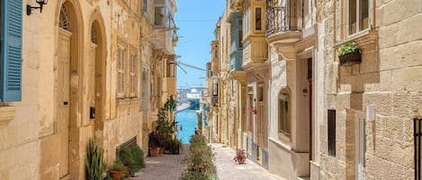 A narrow street with buildings on either side, a canal in the distance, and potted plants along the walkway.