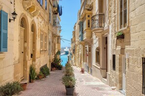 A narrow street with buildings on either side, a canal in the distance, and potted plants along the walkway. - Dar Vitorja (L-Isla)