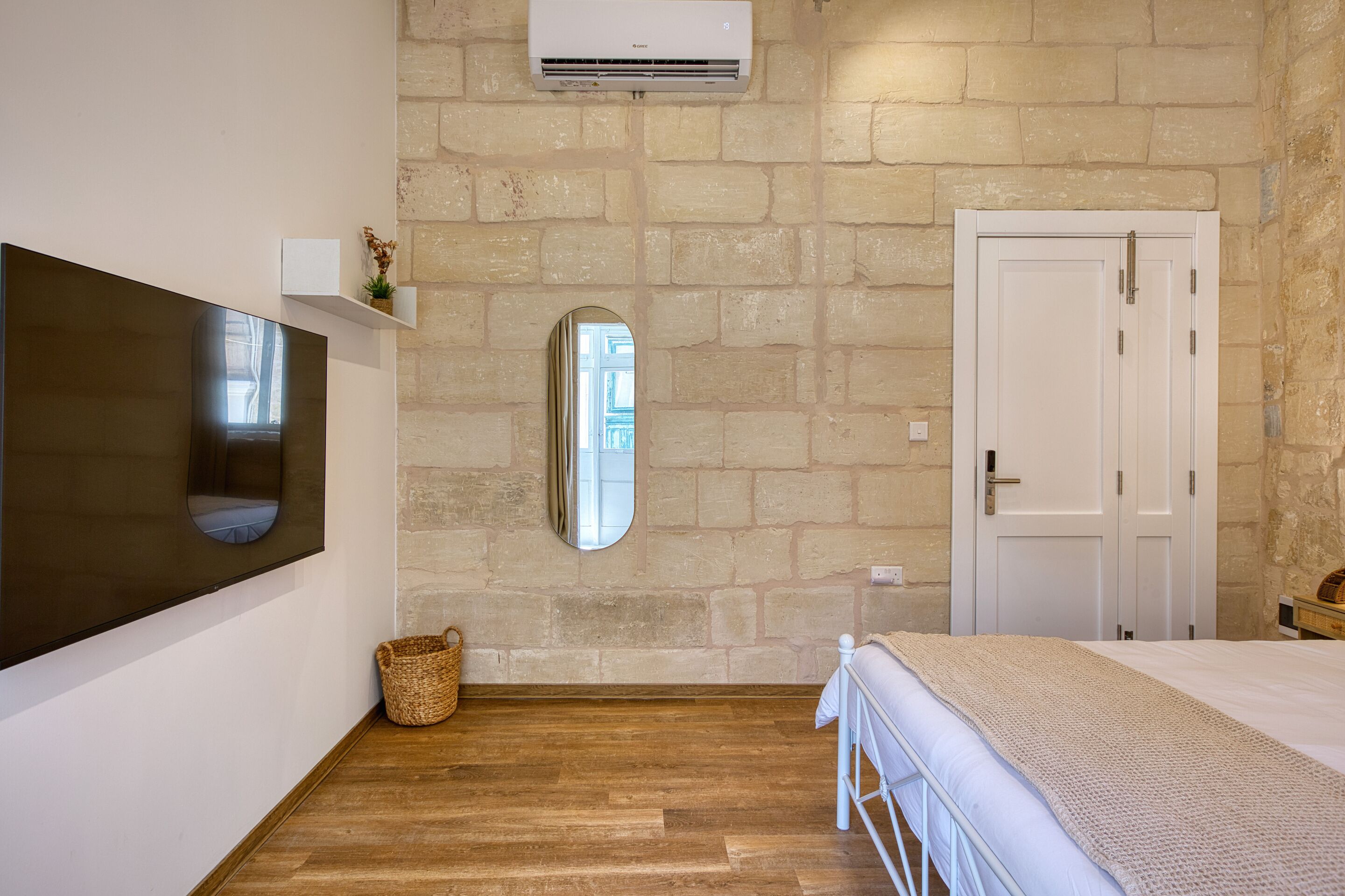 A bedroom with a stone wall, a mounted air conditioner, a bed with a white headboard, a bedside table, and a woven basket.