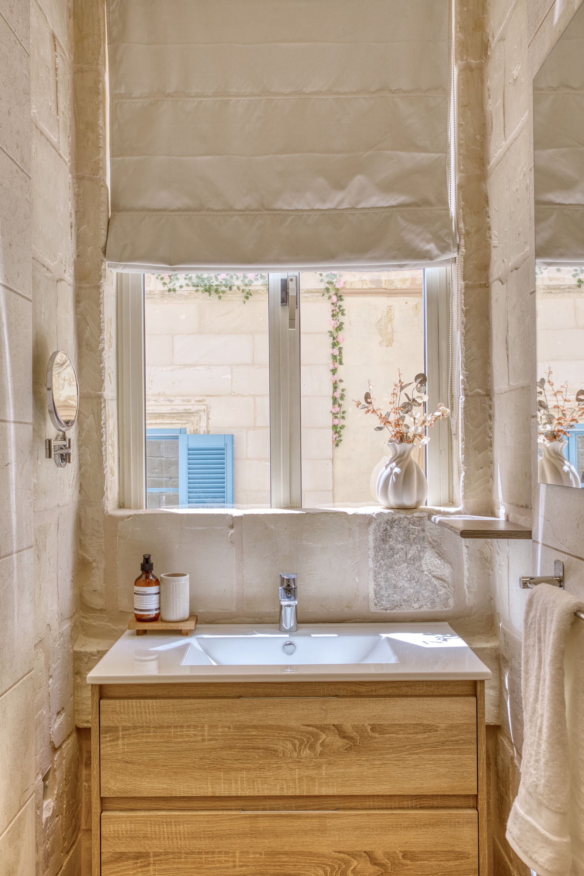 Un baño con lavamanos blanco, mueble de madera y ventana con persianas azules.