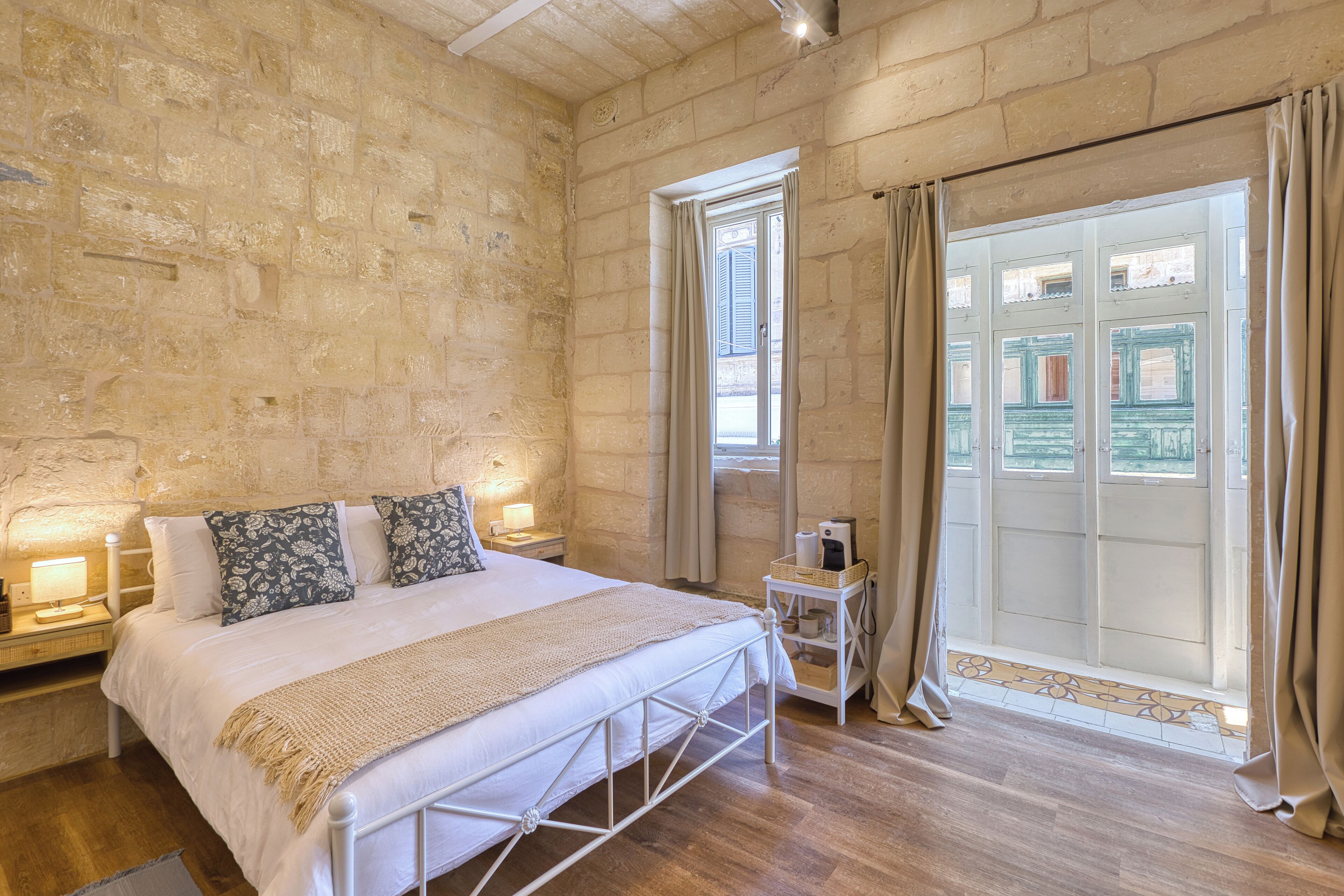 A bedroom with a stone wall, a bed with a white headboard, a bedside table with a lamp, and a window with curtains.