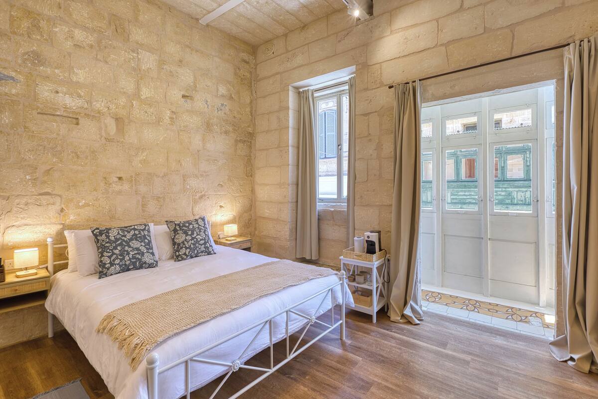 A bedroom with a stone wall, a bed with a white headboard, a bedside table with a lamp, and a window with curtains.