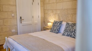 A bedroom with a bed, a nightstand with a lamp, a door, and stone walls. - Dar Vitorja (Senglea)
