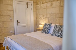 A bedroom with a bed, a nightstand with a lamp, a door, and stone walls.