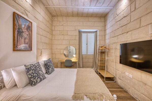 A bedroom with a bed, a desk, a chair, a TV, and a window with blinds.