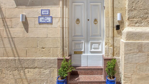 A white double door with a decorative metal crest, flanked by two potted plants and a wall-mounted light fixture. - Dar Vitorja (Senglea)
