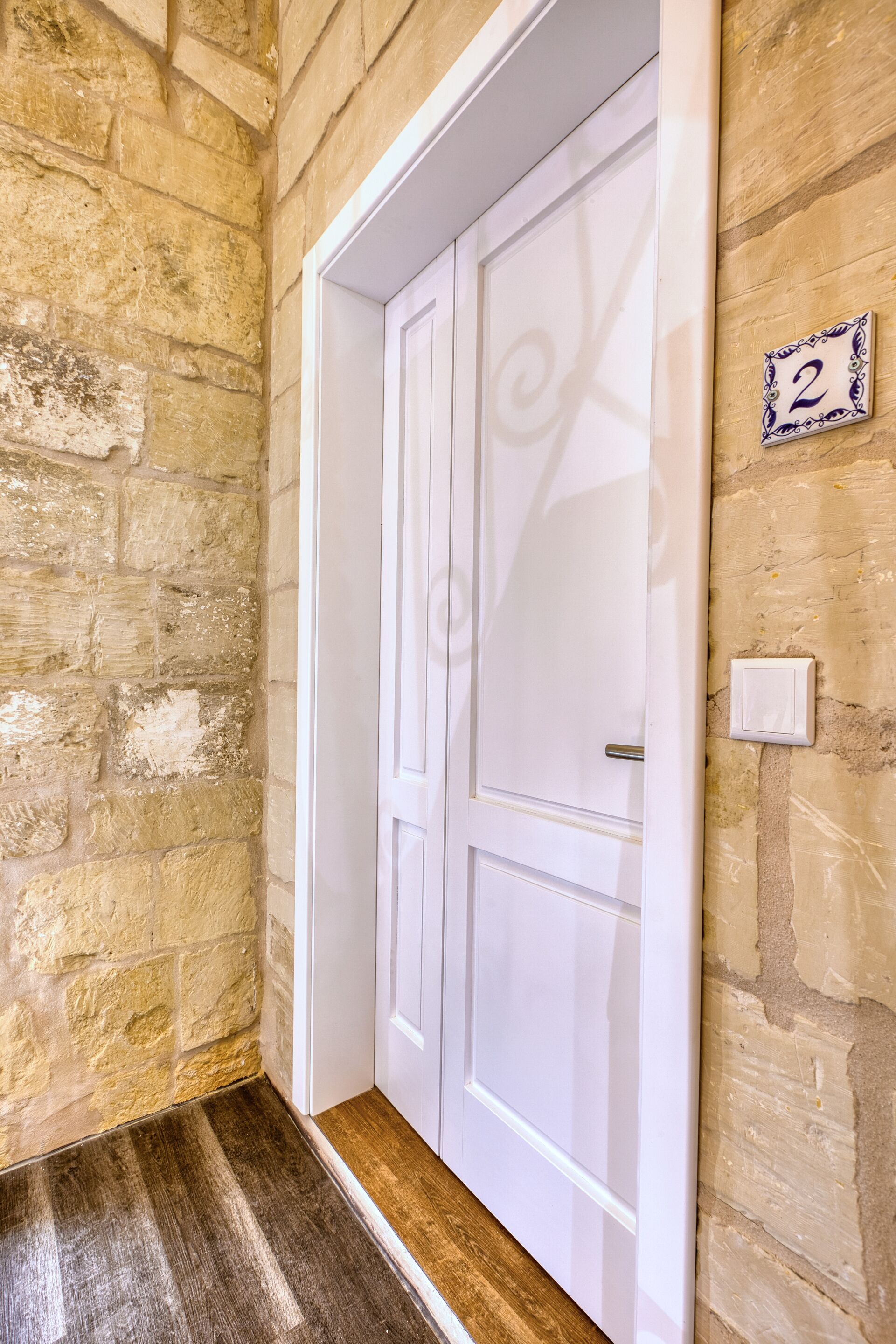 A white door with a decorative design, a number plaque, and a stone wall.