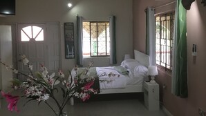 Een slaapkamer met een bed, nachtkastje, lamp, airconditioning, raam met tralies en een plafondventilator.