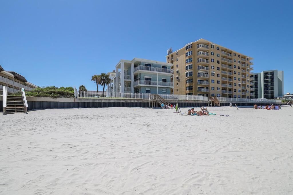 A beachfront hotel with multiple floors, a sandy beach in the foreground, and a clear blue sky.