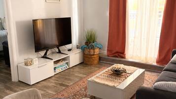 Una sala de estar con televisor de pantalla plana, un sofá, una mesa de centro y un ventanal con cortinas.