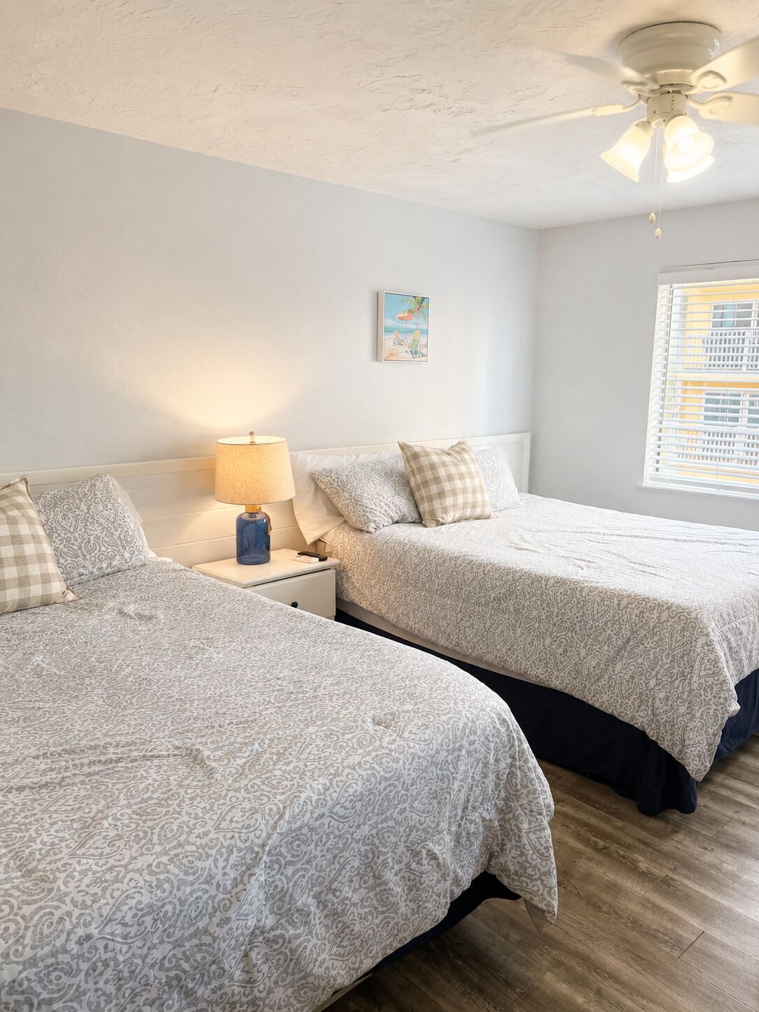 A bedroom with two beds, a nightstand with a lamp, a ceiling fan, and a window with blinds.