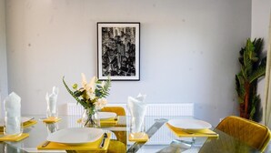 A dining area with a glass table, yellow chairs, white plates, and a framed black and white cityscape picture on the wall. - Joyden's Wood 2-bed Apt, Sleeps 5 With Parking (Dartford)