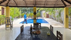 Un espace extérieur abrité, équipé d’une table et de chaises, d’un ventilateur de plafond, et avec une piscine en arrière-plan.