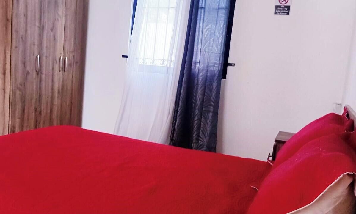Una habitaci ón con un armario de madera, una cama con colcha roja y una ventana con cortinas translúcidas.