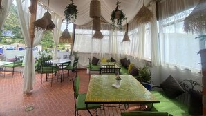 Katettu ulkoalue, jossa on pöytiä ja tuoleja, amppeleissa roikkuvia kasveja ja näkymä ulos.