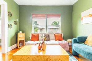 A living room with a sofa, a coffee table, and a blue chair. There is a window with blinds, a plant on a side table, and a wall clock. - Relax & Recharge • King Bed Retreat • Sleeps 4 (Dallas)