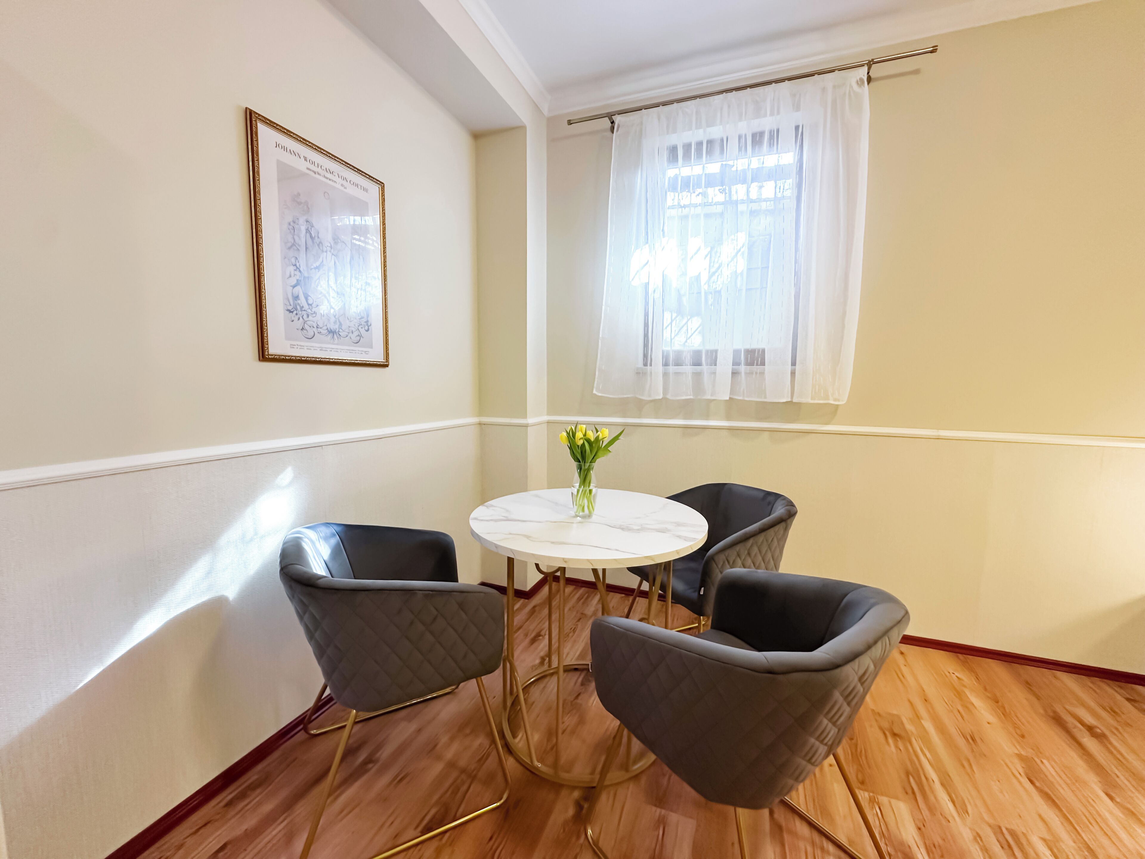 Ein Zimmer mit einem runden Tisch, zwei Stühlen, einem gerahmten Bild an der Wand und einem Fenster mit durchsichtigen Vorhängen.
