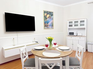 A dining area with a round table, white chairs, a TV mounted on the wall, a poster, and a kitchen area with cabinets and appliances. - Sophia Neighborhood - Spacious 92-square-meter basement apartment near Goethepark (Weimar)