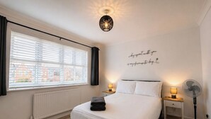 A bedroom with a bed, bedside tables, a wall-mounted light fixture, a standing fan, and a window with blinds.
