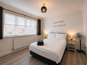 A bedroom with a bed, bedside lamps, a standing fan, a radiator, and a window with blinds.