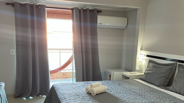 A hotel room with a bed, a nightstand, a lamp, a ceiling fan, and a window with curtains. - Two-room suite equipped with air conditioning in the center of Arraial do Cabo, RJ. (Arraial do Cabo)