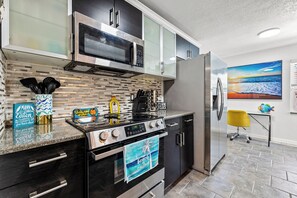 Una cucina moderna con piano cottura, microonde e frigorifero in acciaio inossidabile, zoccolo a mosaico e un asciugamano a tema spiaggia appeso alla porta del forno.