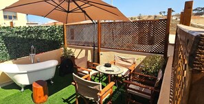 Eine Terrasse mit Badewanne, Holzstühlen, einem Tisch und einem Sonnenschirm.