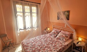 Ein Schlafzimmer mit Bett, Nachttisch mit Lampe, Sessel, Fenster mit Vorhängen und einem Wandbild.