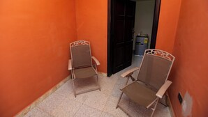Un pasillo estrecho con dos sillas metálicas, una puerta oscura y un pequeño basurero.