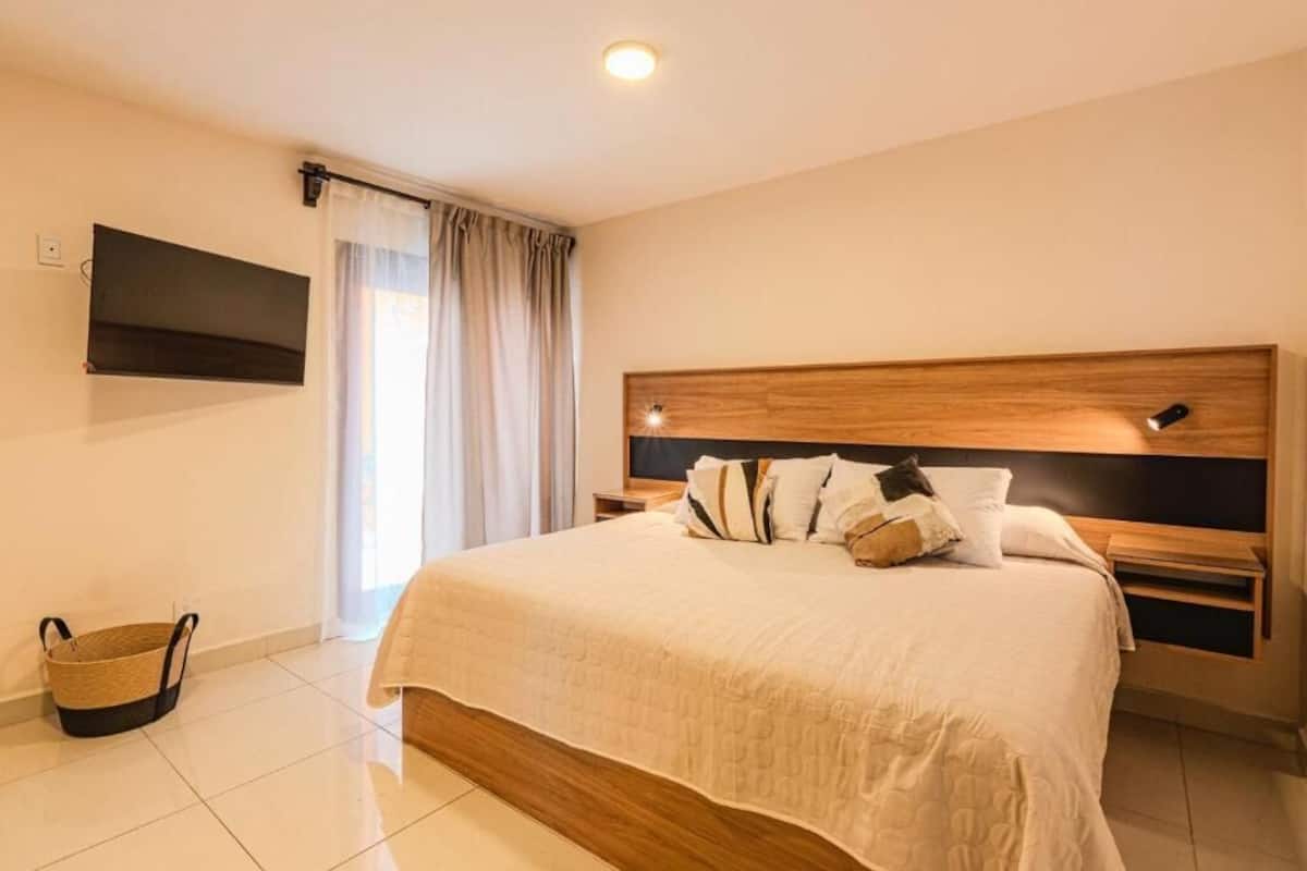 A hotel room with a large bed, a flat-screen TV mounted on the wall, a wooden headboard, and a window with curtains.