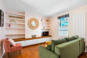 Ein modernes Wohnzimmer mit einem grünen Sofa, einem rosafarbenen Sessel, Holzregalen, einem Netflix-Fernseher und einem Fenster mit Jalousien.