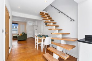 Ein modernes Interieur mit einer Holztreppe, einer Essecke mit weißen Stühlen und einer Küche mit schwarzer Arbeitsplatte.