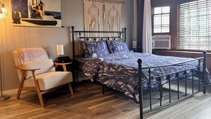 A bedroom with a bed, a chair, a desk, a macrame wall hanging, and a painting.