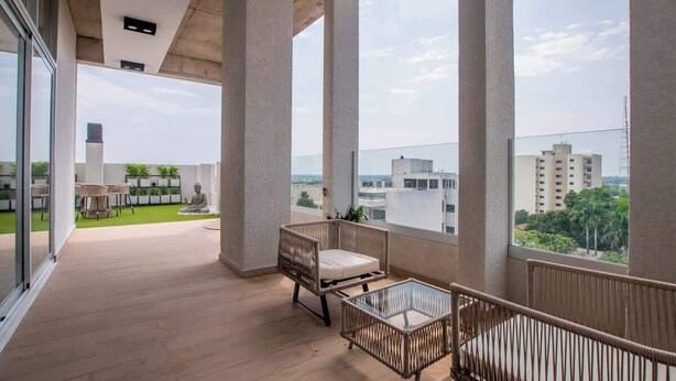 A balcony with a wicker chair, a glass table, and a view of buildings and greenery. - Corporate Stays CorAr 1L (Asunción)