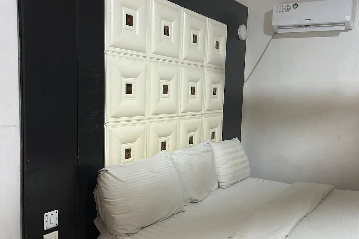 A hotel room with a white bed, a black nightstand with a phone, a white headboard, and an air conditioning unit on the wall.